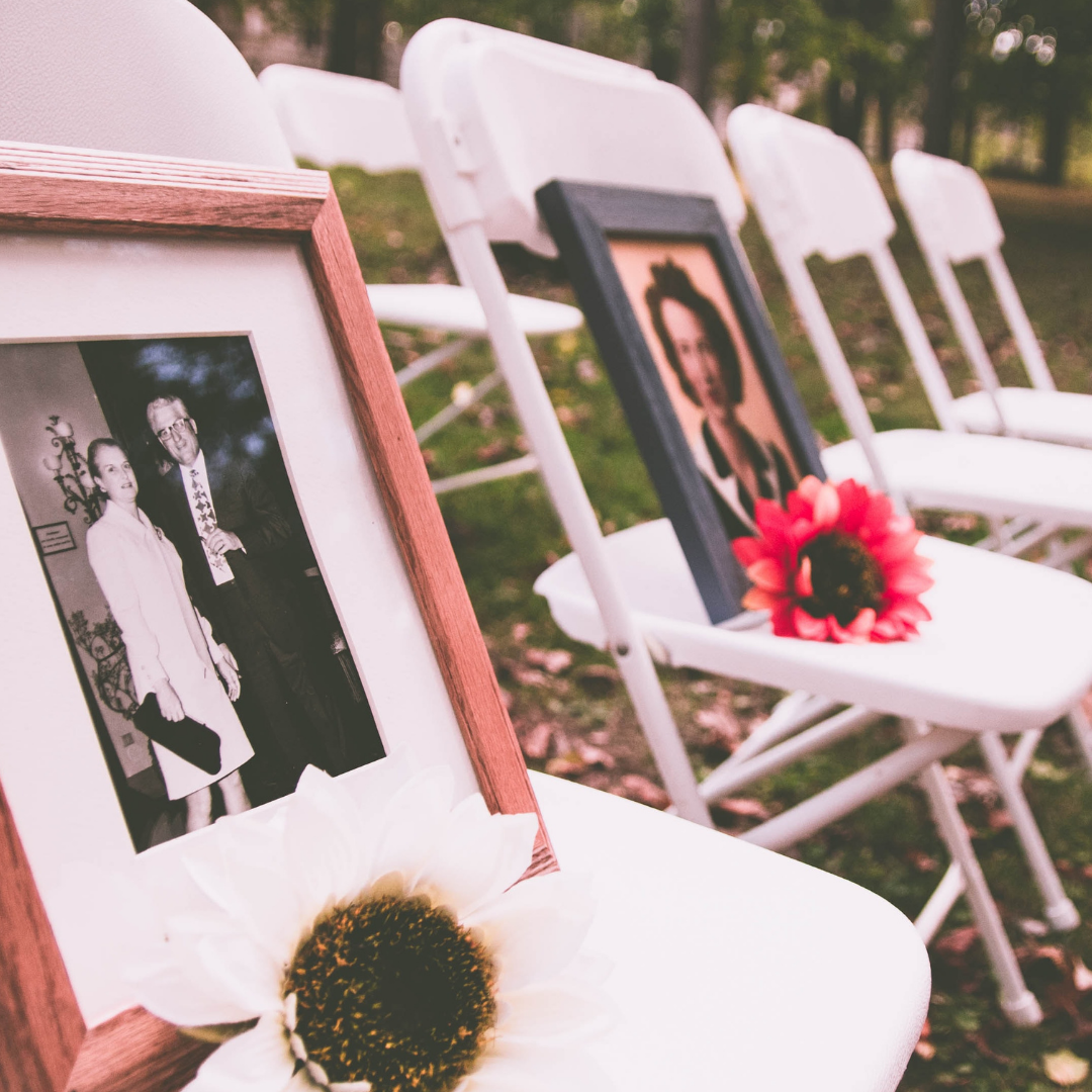 White chairs in a line, two chairs have flowers and picture frames of loved ones on them.