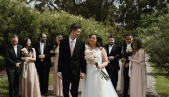 bridal party in pink and black suits standing behind bride and groom looking at each other